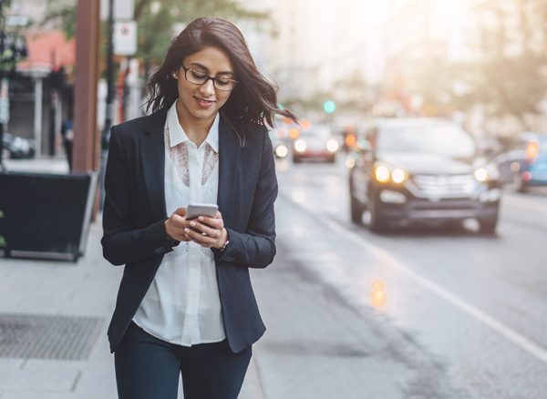 Business woman walking down street while looking at Corporate Travel Management app on mobile phone